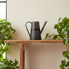 Black watering can on a wooden table with plants around by Aurelis Design Studio