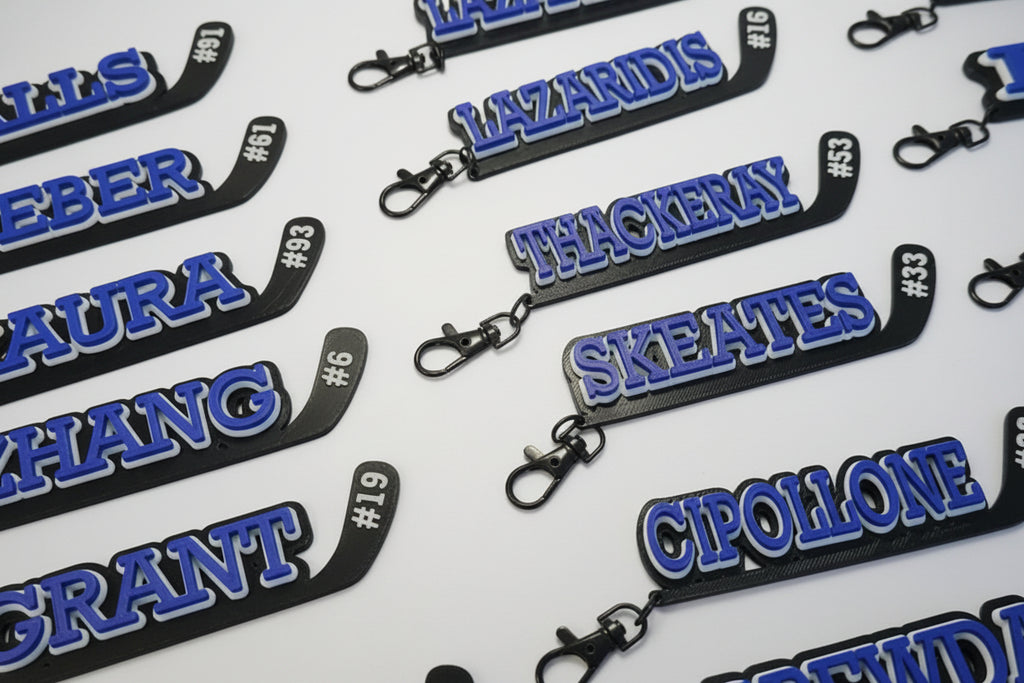 Multiple hockey stick keychains with player names and numbers printed on them in 3 layers with the base in black, the middle layer is white and the name and number is in blue