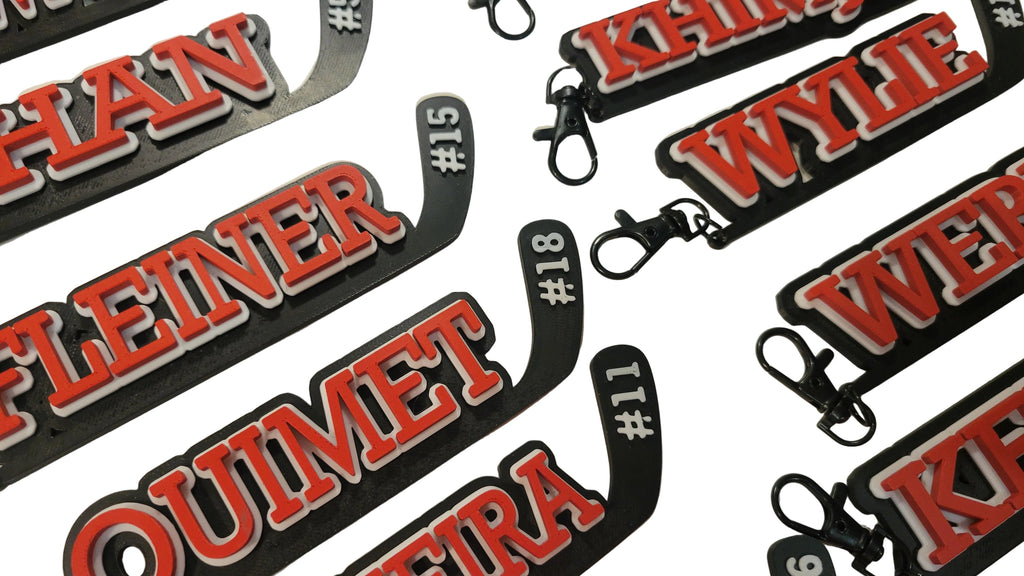 Multiple hockey stick keychains with player names and numbers printed on them in 3 layers with the base in black, the middle layer is white and the name and number is in red