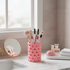 Pink heart-patterned makeup brush holder with brushes on a bathroom counter by Aurelis Design Studio.